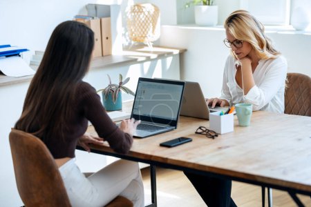 Téléchargez les photos : Prise de vue de deux élégantes femmes d'affaires attrayantes parlant tout en travaillant ensemble avec un ordinateur portable dans une start-up moderne - en image libre de droit