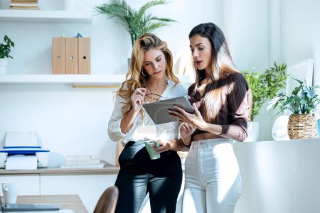 Téléchargez les photos : Prise de vue de deux jeunes femmes d'affaires attrayantes travaillant avec la tablette numérique tout en parlant dans un bureau moderne - en image libre de droit