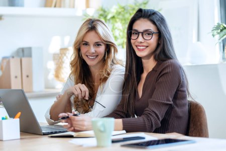 Téléchargez les photos : Prise de vue de deux élégantes femmes d'affaires attrayantes regardant la caméra tout en travaillant avec un ordinateur portable dans une start-up moderne - en image libre de droit