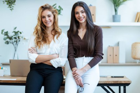 Foto de Fotografía de dos elegantes empresarias atractivas que trabajan mientras miran la cámara en una startup moderna - Imagen libre de derechos