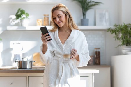 Téléchargez les photos : Tourné de jeune femme souriante profitant d'une tasse de café tout en utilisant son téléphone portable dans le salon à la maison. - en image libre de droit