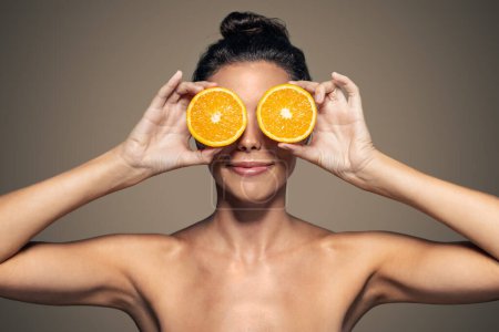 Foto de Retrato de mujer atractiva divertida sosteniendo rebanadas de naranja en su cara y mirando a la cámara en negro marrón aislado - Imagen libre de derechos