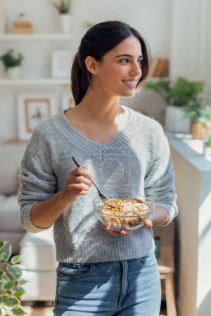 Foto de Foto de una joven sonriente comiendo un tazón de cereales en el desayuno en la sala de estar en casa. - Imagen libre de derechos