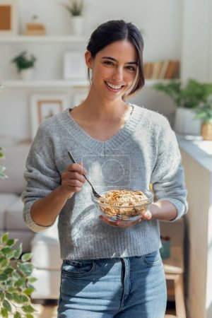 Téléchargez les photos : Prise de vue d'une jeune femme souriante mangeant un bol de céréales au petit déjeuner en regardant la caméra dans le salon à la maison. - en image libre de droit