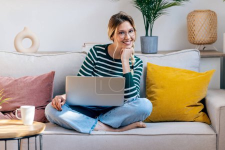 Téléchargez les photos : Tourné de jolie jeune femme travaillant avec son ordinateur portable tout en étant assis sur le canapé à la maison. - en image libre de droit