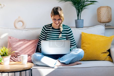 Aufnahme einer schönen jungen Frau, die mit ihrem Laptop arbeitet, während sie zu Hause auf einer Couch sitzt.