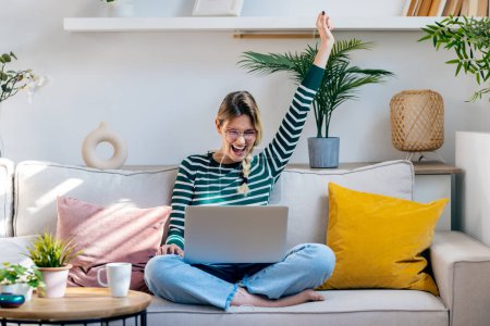 Photo for Shot of pretty motivated woman celebrating something while working with laptop sitting on a couch at home - Royalty Free Image