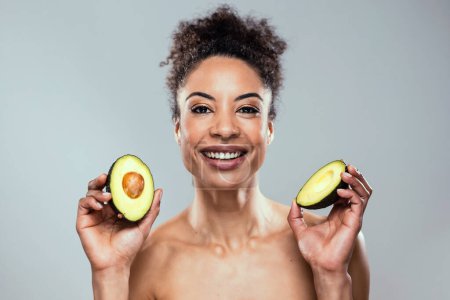 Photo for Portrait of funny attractive woman holding avocado slices at her face and looking at camera on isolated brown blackground - Royalty Free Image