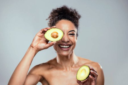 Photo for Portrait of funny attractive woman holding avocado slices at her face and looking at camera on isolated brown blackground - Royalty Free Image