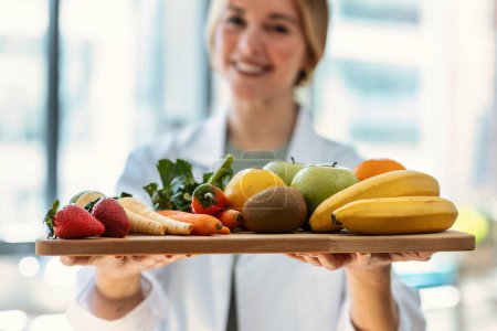 Foto de Primer plano de hermosa nutricionista sonriente sosteniendo mesa de madera con frutas y verduras frescas en una consulta médica - Imagen libre de derechos