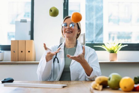 Foto de Foto de hermosa joven nutricionista divirtiéndose mientras hace malabares con una manzana y una naranja en una consulta nutricionista - Imagen libre de derechos