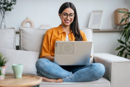Fotografía de una joven sonriente trabajando con su portátil mientras está sentada en un sofá en casa