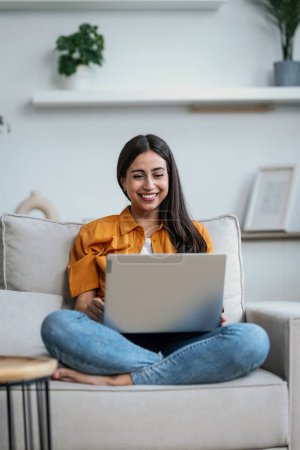 Shot of smiling young woman working with her laptop while sitting on a couch at home