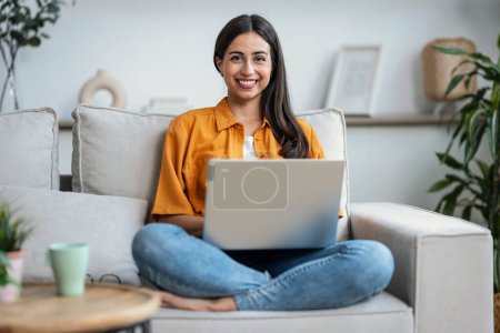 Foto de Fotografía de una joven sonriente trabajando con su portátil mientras está sentada en un sofá en casa - Imagen libre de derechos