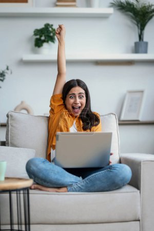 Foto de Tiro de mujer feliz joven celebrando algo mientras trabaja con el ordenador portátil sentado en un sofá en casa - Imagen libre de derechos