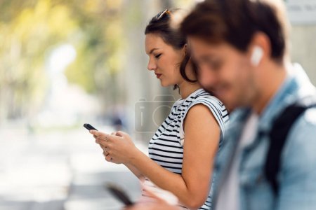 Téléchargez les photos : Prise de vue d'une femme joyeuse utilisant son téléphone portable en attendant le tramway assis à la gare - en image libre de droit