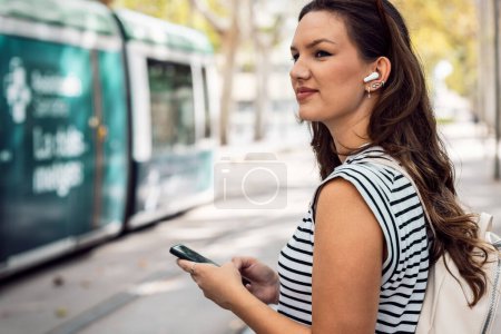 Shot of confident woman listening to music with her mobile phone while waiting for the tram at the station
