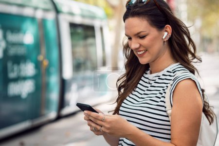 Foto de mujer segura escuchando música con su teléfono móvil mientras espera el tranvía en la estación
