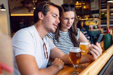 Téléchargez les photos : Tourné de joli jeune couple buvant de la bière tout en utilisant smartphone le bar - en image libre de droit