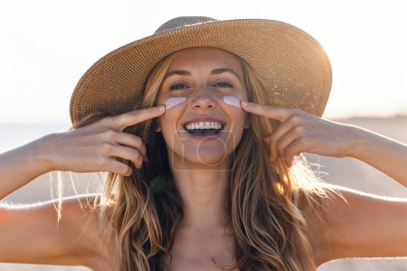 Porträt einer schönen lächelnden Frau, die Sonnencreme auf ihr Gesicht aufträgt, während sie in die Kamera am Strand blickt.