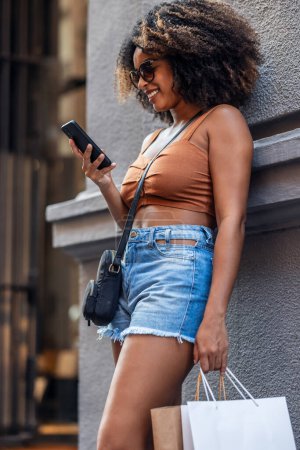 Fotografía de una mujer feliz usando un smartphone mientras llevaba bolsas de compras apoyadas contra la pared de la ciudad