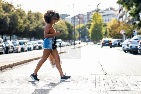 Téléchargez les photos : Prise de vue de belle femme utilisant un smartphone lors de la traversée d'un passage de zèbre dans la ville - en image libre de droit