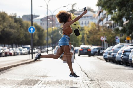 Foto de Tiro de mujer loca divertida saltando mientras sostiene el teléfono inteligente cruzando la calle. - Imagen libre de derechos