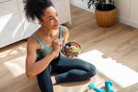 Tiro de mujer atlética comiendo un tazón de fruta saludable mientras se sienta en el suelo en la cocina en casa