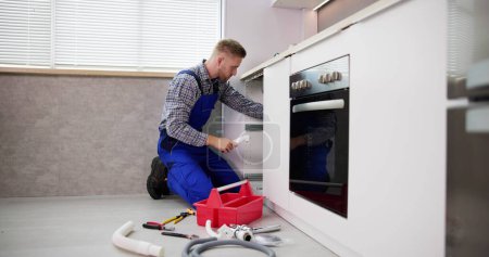 Téléchargez les photos : L'homme regardant le plombier masculin nettoyant le tuyau d'évier bloqué avec le câble vidé dans la cuisine - en image libre de droit