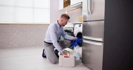 Téléchargez les photos : Jeune homme chargement des vêtements dans la machine à laver dans la cuisine - en image libre de droit