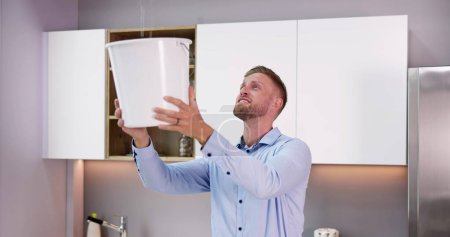Téléchargez les photos : Homme inquiet tenant seau tandis que les gouttelettes d'eau fuient du plafond dans la cuisine - en image libre de droit