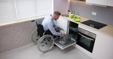 Téléchargez les photos : Personne handicapée en fauteuil roulant utilisant un lave-vaisselle dans la cuisine - en image libre de droit