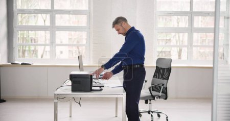 Photo for Irritated Businessman Looking At Paper Stuck In Printer At Office - Royalty Free Image