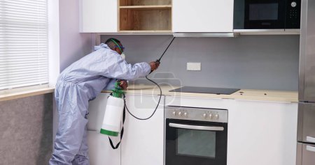 Photo for Man Showing At Pest Control Worker Spraying Insecticide On Shelf Of Domestic Kitchen - Royalty Free Image