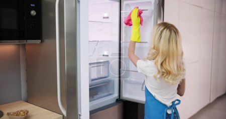 Photo for Young Smiling Professional Cleaning Service Woman Cleaning Refrigerator In Kitchen - Royalty Free Image