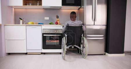 Téléchargez les photos : Jeune homme handicapé assis sur fauteuil roulant préparant la nourriture dans la cuisine - en image libre de droit