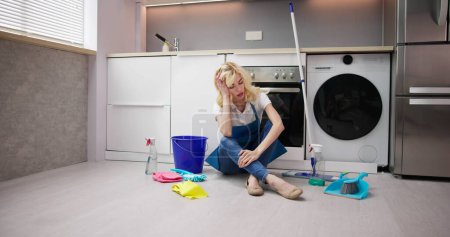 Téléchargez les photos : Frustrée jeune femme assise sur le plancher de la cuisine et regardant les produits de nettoyage à la maison - en image libre de droit