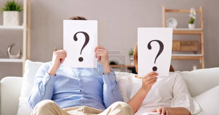 Photo for Couple Sitting On Sofa Holding Question Mark Sign In Front Of Face - Royalty Free Image