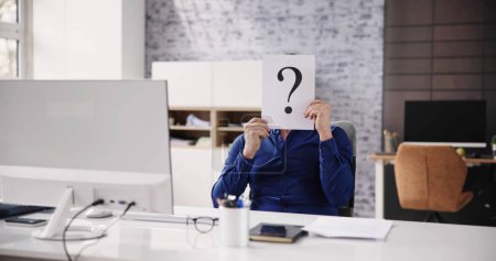 Photo for Businessman Holding Paper With Question Mark In Front Of Face At Desk In Office - Royalty Free Image