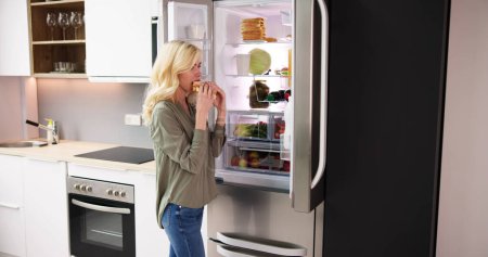 Téléchargez les photos : Sandwich au fromage affamé femme manger près d'un réfrigérateur ouvert - en image libre de droit