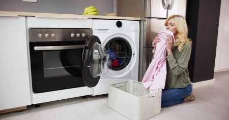 Photo for Woman Smelling Clean Clothes Near The Electronic Washer At Laundry Room - Royalty Free Image