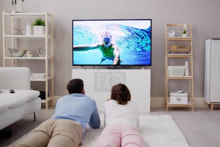 Foto de Joven feliz pareja acostado en alfombra viendo película en televisión en casa - Imagen libre de derechos