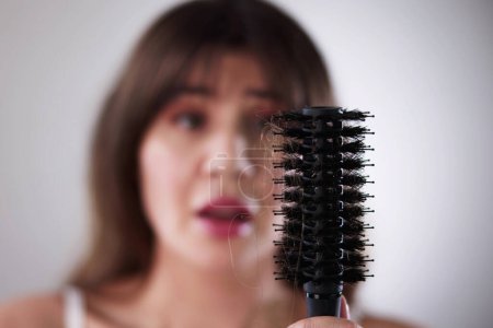 Foto de Mujer joven en albornoz sosteniendo peine mirando la pérdida de cabello en casa - Imagen libre de derechos