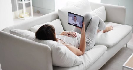 Téléchargez les photos : Jeune femme enceinte visioconférence avec médecin sur tablette à la maison - en image libre de droit