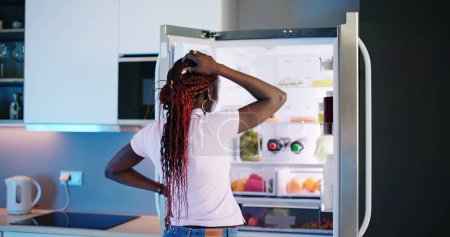 Photo for Young Woman Looking At Food In Open Refrigerator - Royalty Free Image