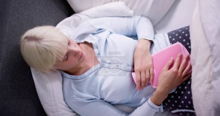 Foto de Mujer joven con botella de agua caliente en el estómago acostado en la cama - Imagen libre de derechos