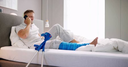 Téléchargez les photos : Jeune homme heureux avec jambe cassée assis sur le canapé parlant sur un téléphone mobile - en image libre de droit
