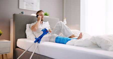 Photo for Happy Young Man With Broken Leg Sitting On Sofa Talking On Mobile Phone - Royalty Free Image