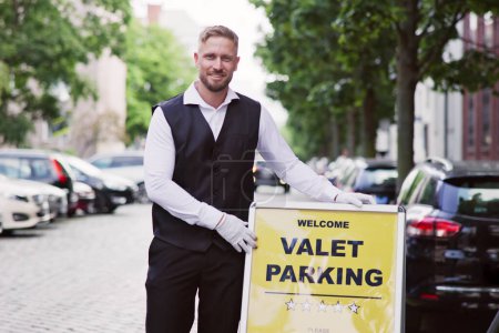 Photo for Portrait Of A Smiling Young Male Valet Standing Near Valet Parking Sign - Royalty Free Image