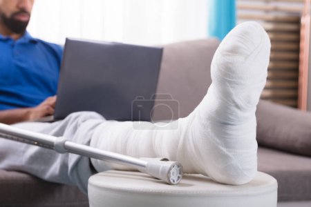 Téléchargez les photos : Homme avec jambe cassée assis sur le canapé en utilisant un ordinateur portable - en image libre de droit